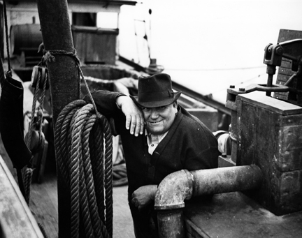 In Southport, Charles "Pete" Joyner identified this man as "Doc" Robinson, pilot of one of the Brunswick Navigation Co.'s other menhaden boats, the John M. Morehead. He recalled him as a “big man, [got] around kind of slow.” He's leaning on the boat's engine room. Next to him is a mechanical pump for draining water out of the fish hole. The line that his right arm rests on raises one end of a purse boat. Behind him is the windlass, run by a chain off the engine, that picks up the two purse boats. Beyond the windless, we can see the bail net and a tank of freshwater behind the pilot house. Photo by Charles A. Farrell. Courtesy, State Archives of North Carolina