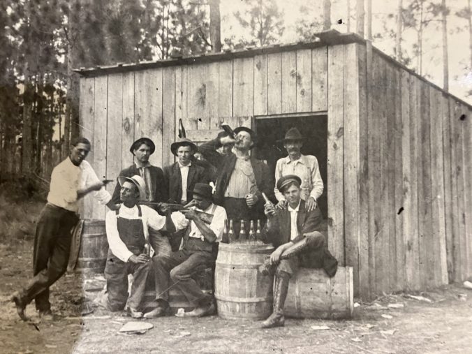 Revelers mugging for the camera at the Makatoka logging camp. One man is holding a pistol, another a rifle, and at least three are holding a bottle. From Waccamaw Lumber Co. Photographs & Journal, Rubenstein Library, Duke University