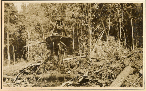 Derrick barge No. 14's orange-peel bucket" cutting off a point on the Scuppernong River, May 1931. An "orange-peel bucket" is a kind of bucket dredge, of which there are 2 kinds. A "clam-shell" bucket has 2 jaws (or shells), while the "orange-peel bucket" used here by derrick barge No. 14 has 3 or 4 jaws. Both work the same way. The bucket hangs from 2 wires or chains hung from the end of the boom and powered by a double cylinder, double drum steam engine. The hoist operator uses those wires to lower the bucket, open the bucket's jaws and draw in earth and vegetation, and close the bucket, and then to raise the bucket, turn the boom, and lower the load onto a scow or, in this case, onto the banks of the Scuppernong. Source: Office of History, HQ, U.S. Army Corps of Engineers
